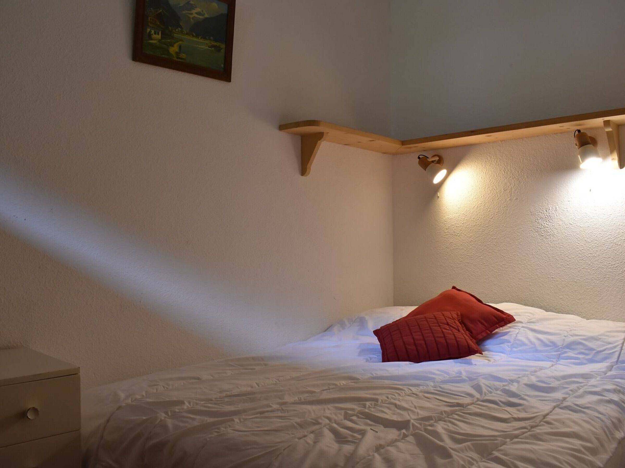 A bedroom with a bed, a nightstand, a wooden shelf with lights, and a painting on the wall.