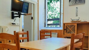 A dining area with a wooden table and chairs, a television mounted on the wall, a window with a view of greenery, and a small cabinet with glass doors. - 2-Room Apartment 4 Pers | Center Cauterets (Cauterets)