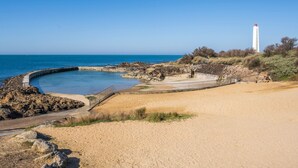 A coastal landscape with a lighthouse, sandy beach, rocky shoreline, and a curved concrete structure. - Charming T3 Apartment near Wild Coast, Pool, and Beaches (Les Sables-d'Olonne)