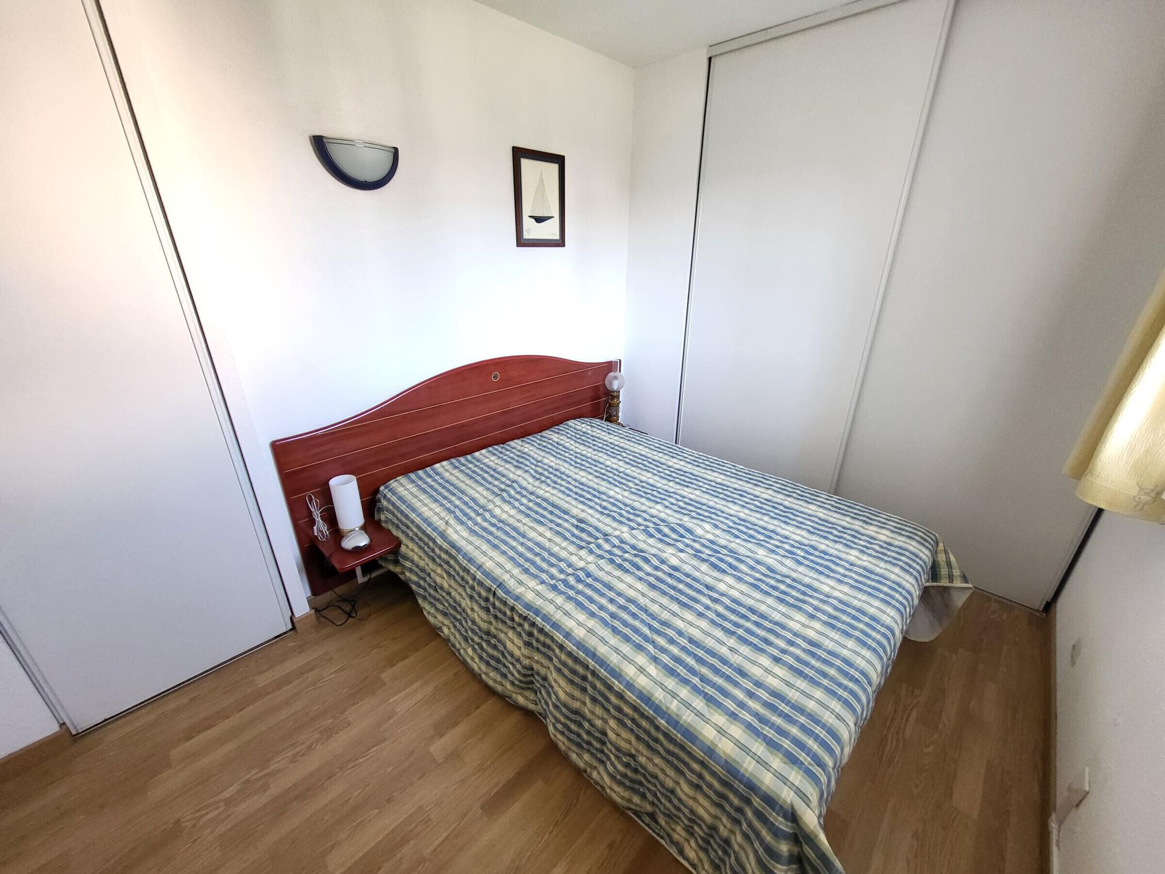A small, neatly arranged bedroom with a wooden floor, a single bed with a striped bedspread, a bedside table with a lamp, and a wall-mounted shelf with a framed picture.