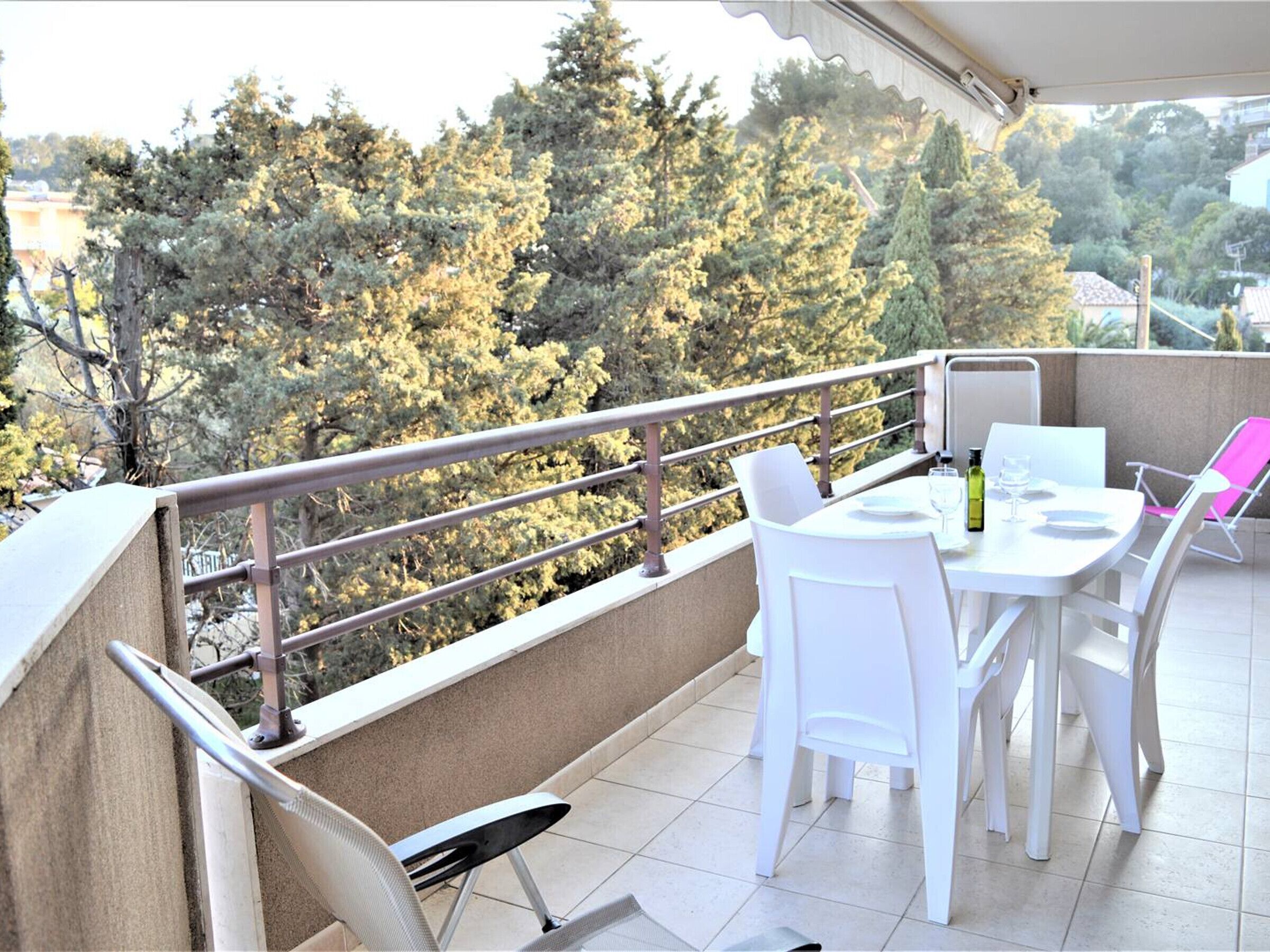 Een balkon met een tafel, stoelen, een hekwerk en uitzicht op bomen en gebouwen.