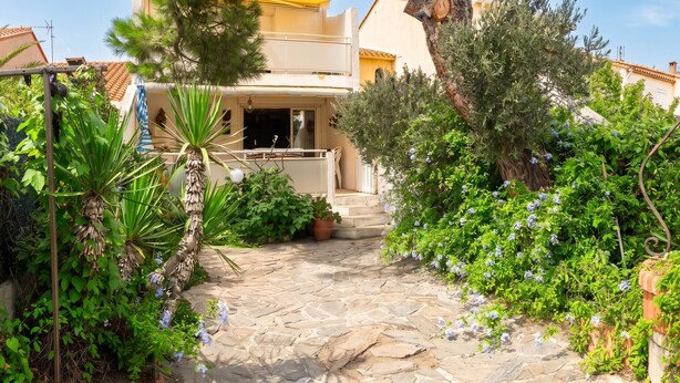 A stone pathway leading to a building with a balcony, surrounded by lush greenery and flowering plants.