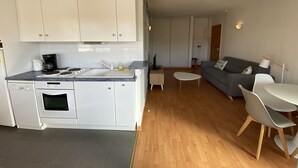 A compact kitchen with white cabinets, a built-in oven, and a coffee maker. There is a dining area with a small table and chairs, and a grey sofa in the living room. - Charming 1-Bed Apt with South-Facing Terrace, Beach Access & Parking in Arcachon (Arcachon)