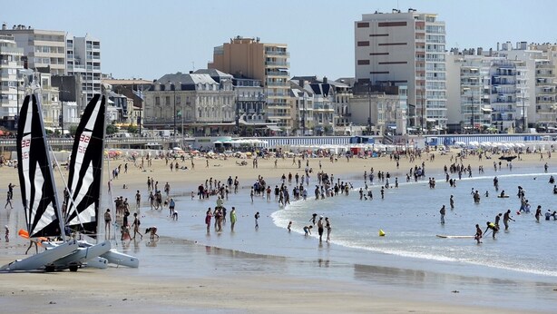 Una playa concurrida, gente, un velero y edificios al fondo.