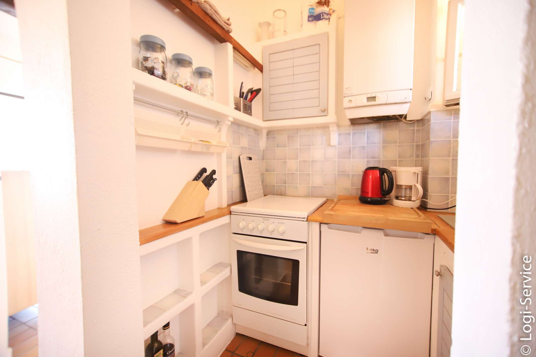 Una cocina compacta con estufa, horno y un refrigerador pequeño. Hay estantes con frascos y utensilios de cocina.
