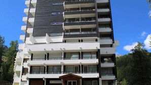 A multi-story hotel with a modern design, featuring balconies on each floor, a central entrance, and a parking area with cars.