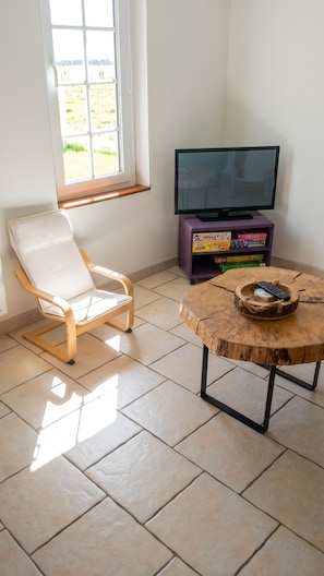 Ein Zimmer mit einem weißen Stuhl, einem Fernseher auf einem violetten Standfuß und einem runden Holztisch mit einer Fernbedienung.
