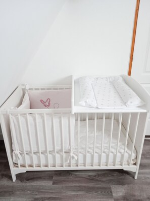 Un berceau blanc, avec un matelas et une couverture, placé contre un mur uni et sur un plancher en bois.