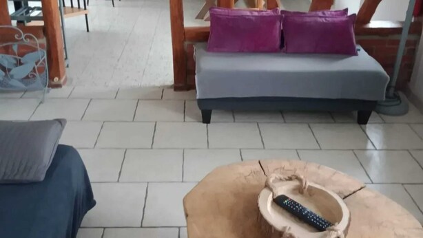 Una habitación con sofá de madera, cama azul y mesa de centro redonda de madera con un control remoto encima.