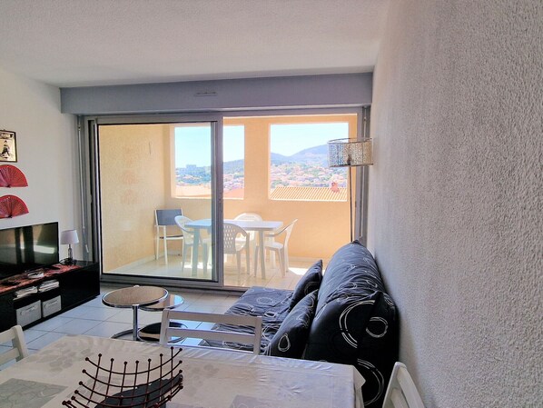 A balcony with a view of a cityscape, a sofa, a TV, and a dining table with chairs.