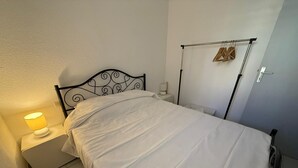 Een netjes opgemaakt bed in een kleine kamer met een nachtkastje, een wandrek om kleding te drogen, en een eenvoudige muur.