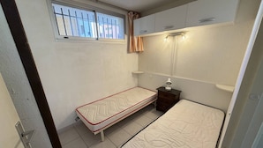 Un dormitorio pequeño con dos camas individuales, una ventana con rejas, una mesita de noche y un armario sobre las camas.