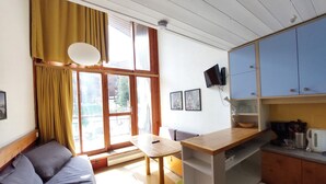 A compact living space with a sofa, a small dining table, a kitchenette with blue cabinets, a TV mounted on the wall, and a window with curtains. - Renovated duplex, pets allowed, for 5 near slopes and shops (Les Arcs 1800)