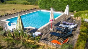 Piscina con sillones de descanso y sombrillas, rodeada de vegetación y colinas.