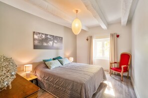 A bedroom with a bed, bedside lamps, a red chair, a window with curtains, and a wall-mounted artwork.