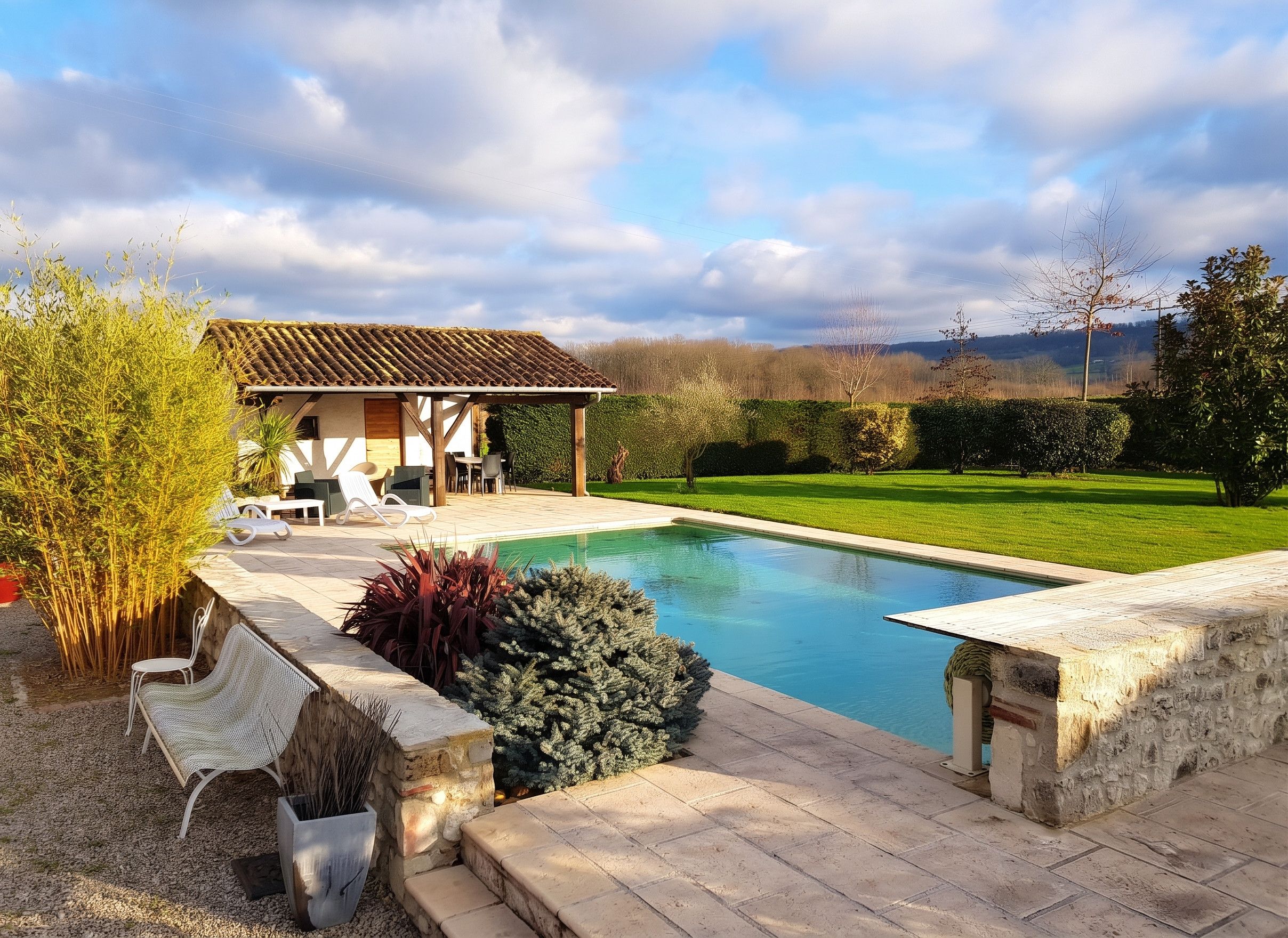 A pool with a stone deck, a covered outdoor seating area, and a view of a green landscape.