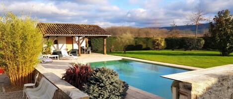A pool with a stone deck, a covered outdoor seating area, and a view of a green landscape.