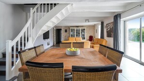 A dining area with a wooden table, wicker chairs, and a fruit bowl. There is a staircase with white railings leading to another room.