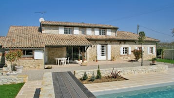 Une maison en pierre à deux étages, avec un toit en tuiles, une piscine et un patio aménagé avec du mobilier d’extérieur.