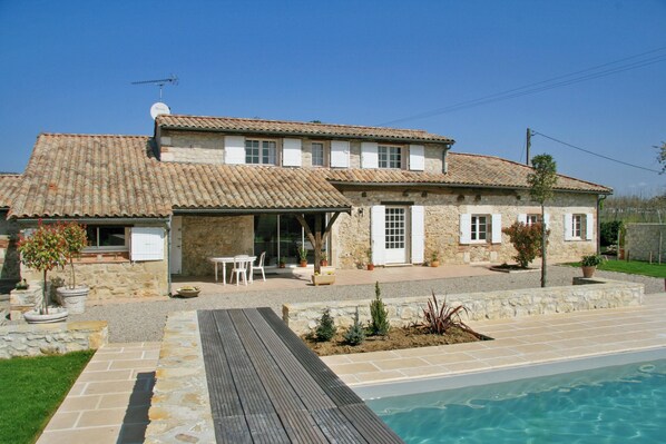 A two-story stone house with a tiled roof, a swimming pool, and a patio area with outdoor furniture.
