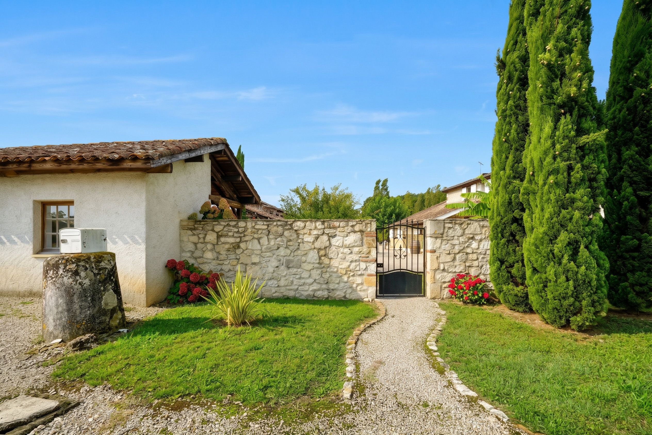 A stone house with a tiled roof, a gravel path leading to a gate, and a well-maintained lawn with red flowers.