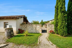 A stone house with a tiled roof, a gravel path leading to a gate, and a well-maintained lawn with red flowers.