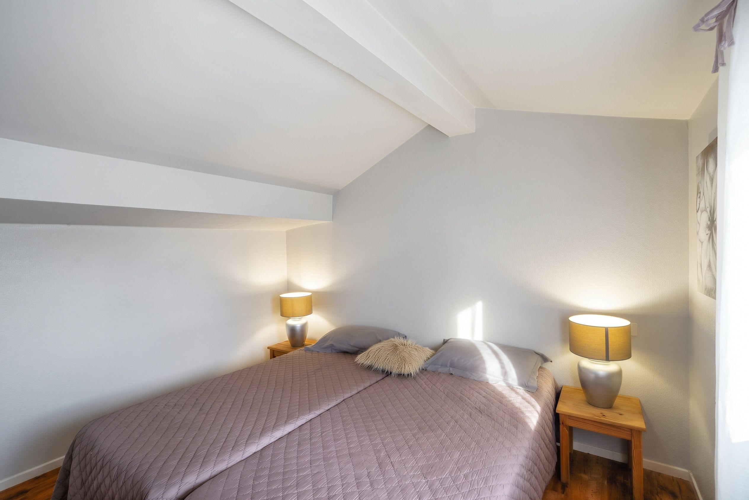 A bedroom with a sloped ceiling, a bed with a quilted cover, two bedside lamps, and a framed picture on the wall.