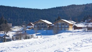 A snow-covered landscape with a row of wooden buildings, likely a resort or lodge, with a forested hill in the background.