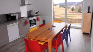 A modern kitchen with a wooden dining table, red and blue chairs, and a view of a rural landscape through large windows.