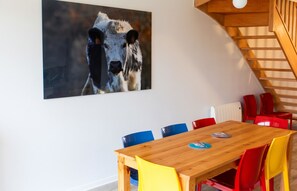 A dining area with a wooden table and colorful chairs, a large painting of a cow on the wall, and a staircase with wooden railings. - Homerez - Apartment in Xonrupt-Longemer (Xonrupt-Longemer)