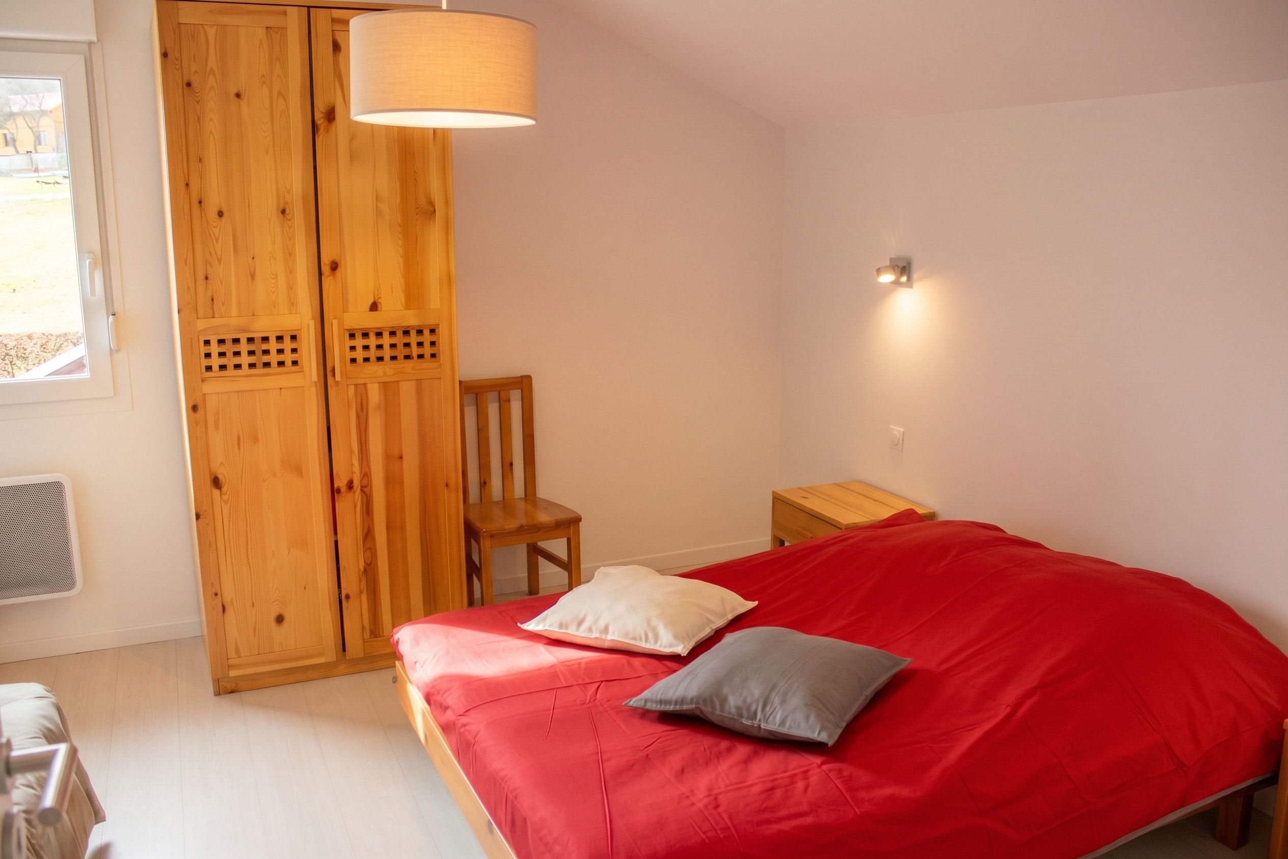 A bedroom with a red bed, a wooden wardrobe, a chair, and a bedside table.