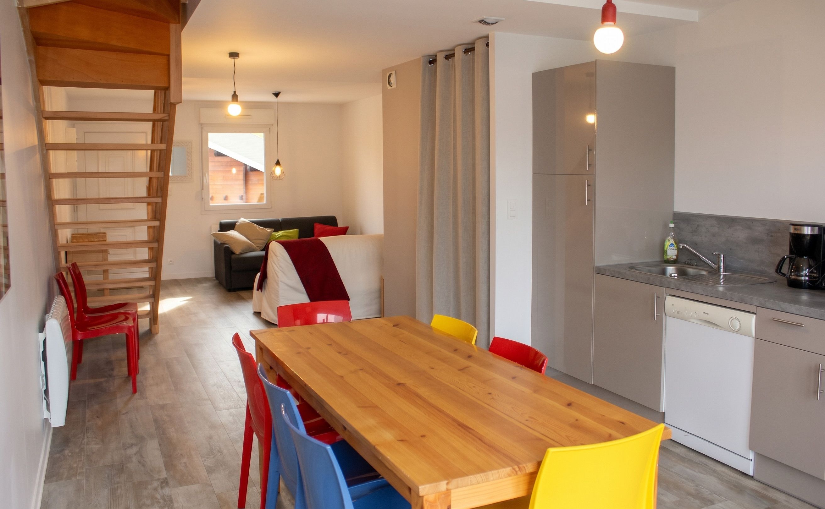 Een moderne keuken met een houten eettafel en kleurrijke stoelen, een ingebouwde oven en een koffiezetapparaat.