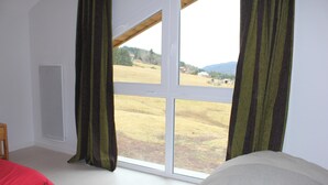 A bedroom with a large window, a bed, and a view of a field and mountains.