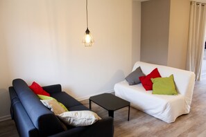 A modern living room with a black sofa, a white sofa with colorful cushions, a small black coffee table, and a hanging light fixture. - Homerez - Apartment in Xonrupt-Longemer (Xonrupt-Longemer)