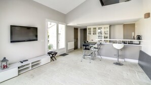 Un appartement moderne doté d’un téléviseur fixé au mur, d’un coin repas avec une table et des chaises, d’une cuisine avec des placards et un plan de travail, et d’une grande fenêtre laissant entrer la lumière naturelle.