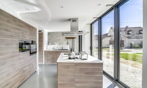 Une cuisine moderne dotée d’un grand îlot central, d’un four encastré et offrant une vue sur la campagne grâce à de grandes fenêtres.