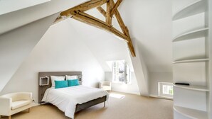 Une chambre avec un plafond en pente, un lit avec une literie blanche et des coussins turquoise, un fauteuil blanc et une bibliothèque intégrée.