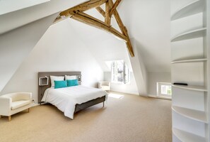 A bedroom with a sloped ceiling, a bed with white bedding and teal pillows, a white armchair, and a built-in shelving unit. - Homerez - Villa with Pool and Garden in Amponville (Amponville)