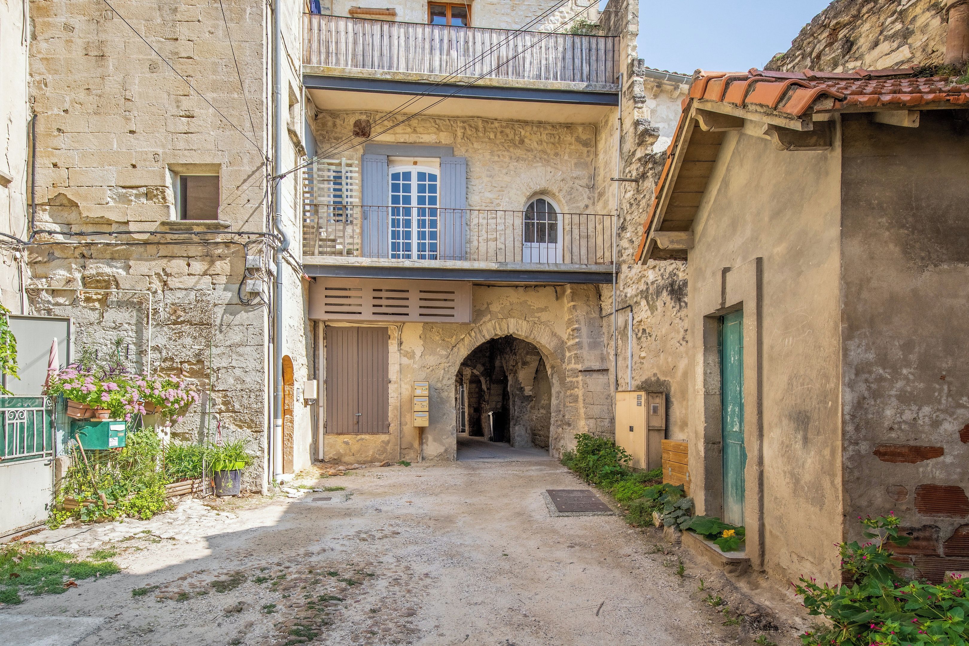 A narrow alleyway with a stone building featuring an arched entrance, a wooden balcony, and a small window with shutters.