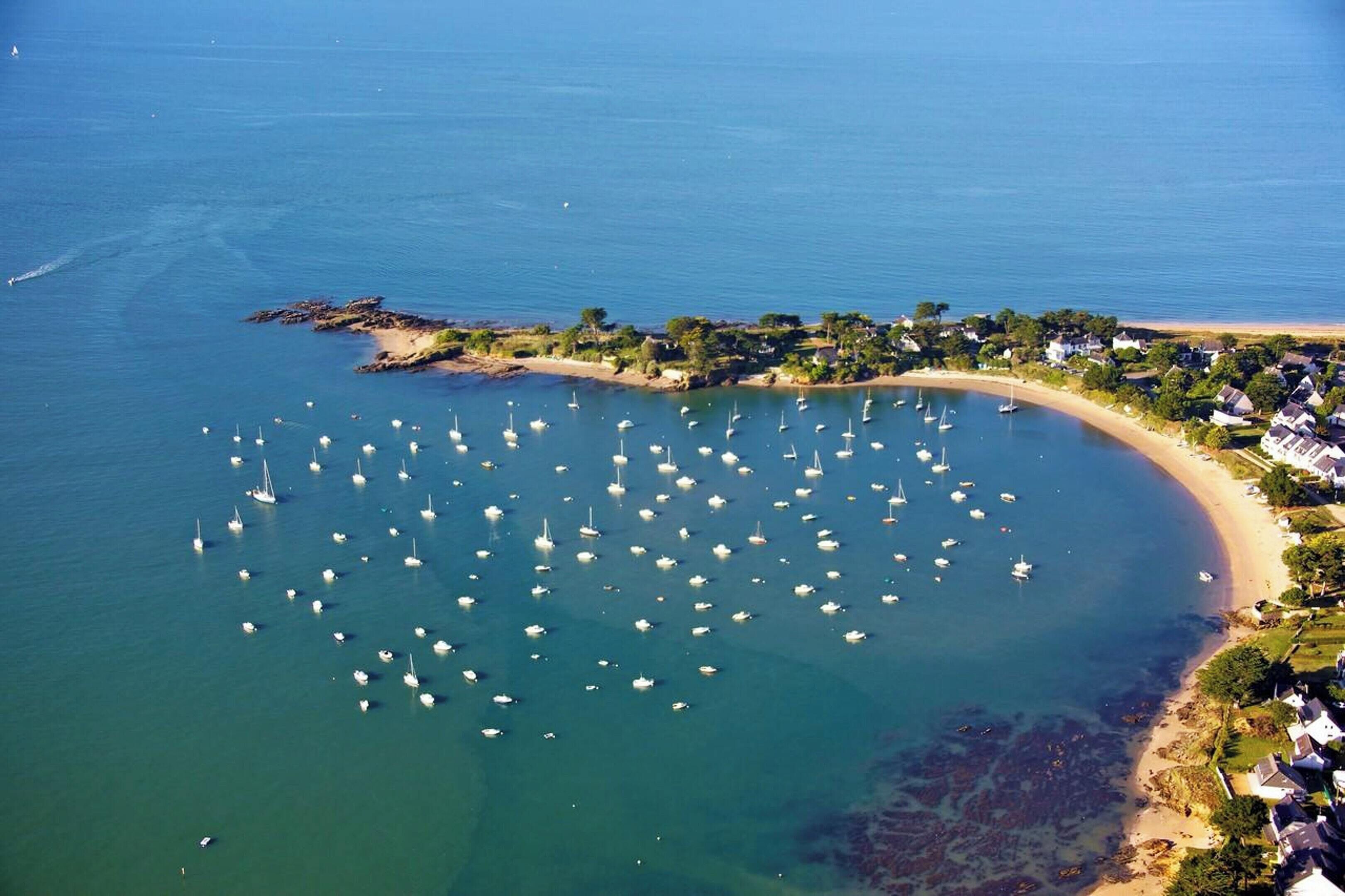An aerial view of a coastal area with numerous boats anchored in a bay, a sandy beach, and a residential area with houses along the shore.