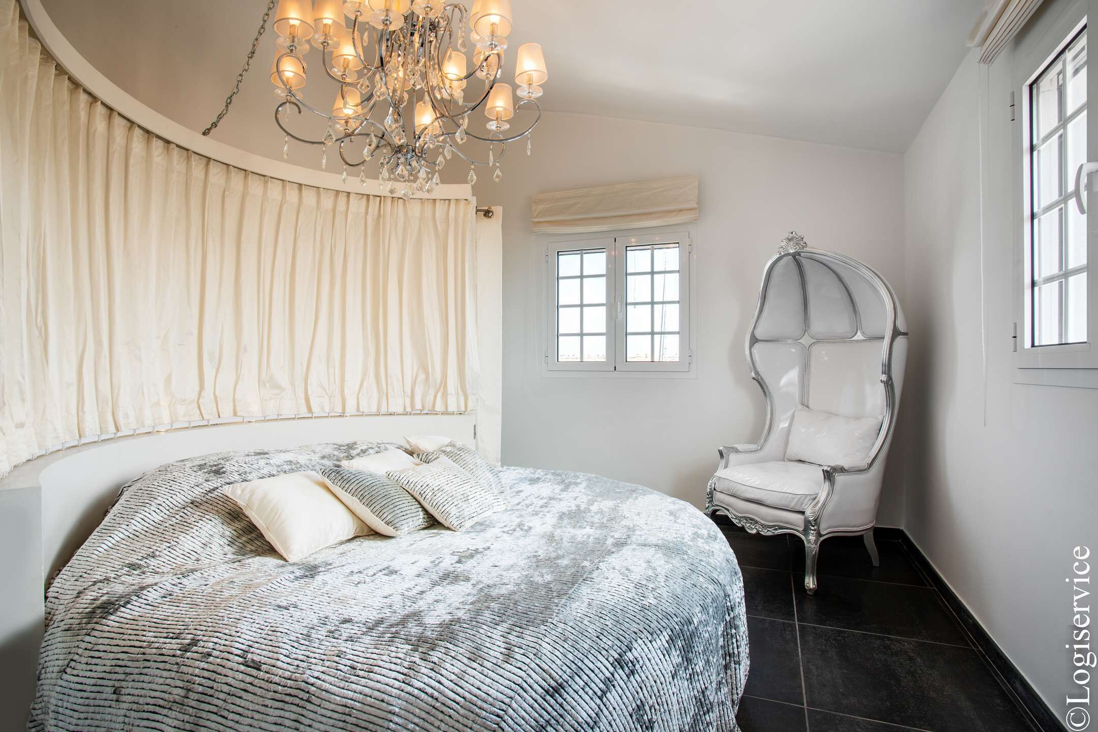 Une chambre à coucher avec un grand lit, un lustre, une chaise blanche et une fenêtre avec des stores.