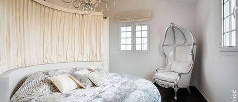 Une chambre à coucher avec un grand lit, un lustre, une chaise blanche et une fenêtre avec des stores.