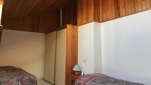 A room with wooden walls and a sloped ceiling, a bed with a patterned bedspread, a bedside table with a lamp, and a wardrobe. - Cozy 2-Bedroom Villa + Mezzanine, Sleeps 6, Steps to Beach, Parking, TV, Washer (Port Leucate)