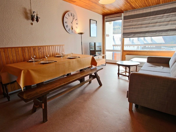 A dining area with a wooden table, benches, and a sofa. There is a clock on the wall and a window with a view of buildings.