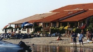 Un edificio costero con techo de tejas, una playa al frente y gente cerca del agua.