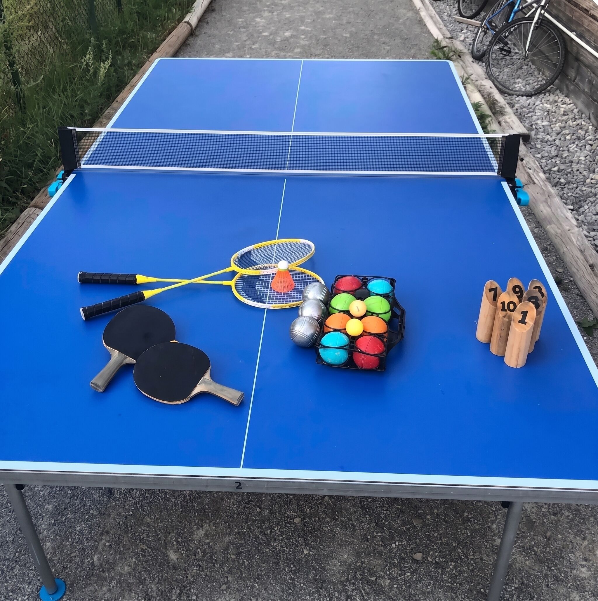 Et blåt bordtennisbord med to bat, en fjerbold, en opbevaringsholder med farvede bolde og nummererede sko.