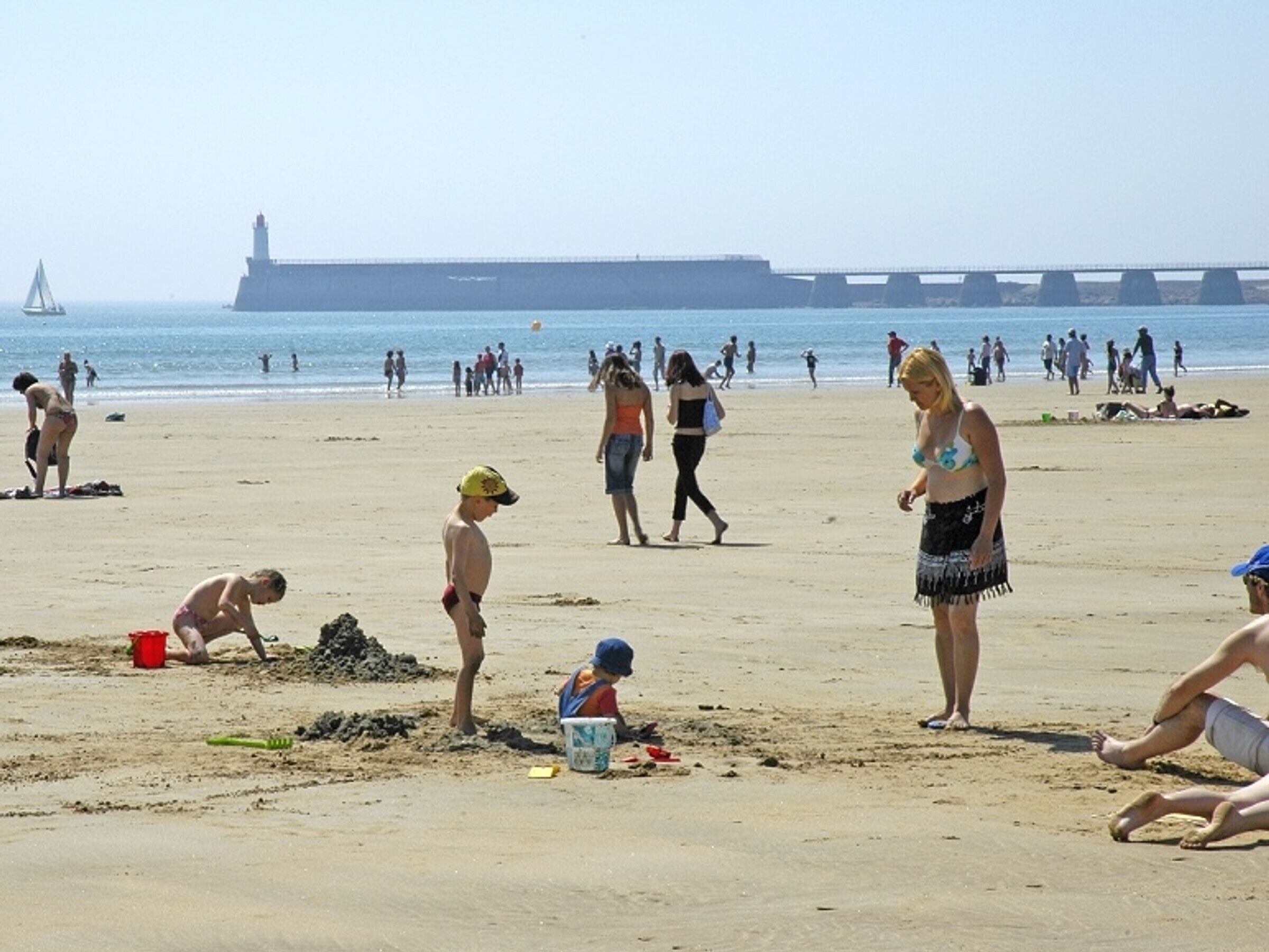 A beach with people, a lighthouse, and a bridge in the background.