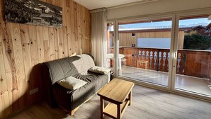 Habitació acollidora amb paret de fusta, un sofà gris, taula de fusta petita i una porta corredissa de vidre que dona a un balcó amb vistes a cases i muntanyes.