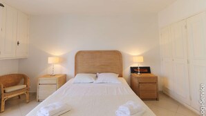 A hotel room with a large bed, two bedside tables with lamps, a wicker chair, and white wardrobes. - Renovated 1 bedroom flat for 2 persons (PORT GRIMAUD)