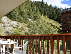 A wooden deck with a table and chairs, overlooking a forested hillside and a building with balconies.